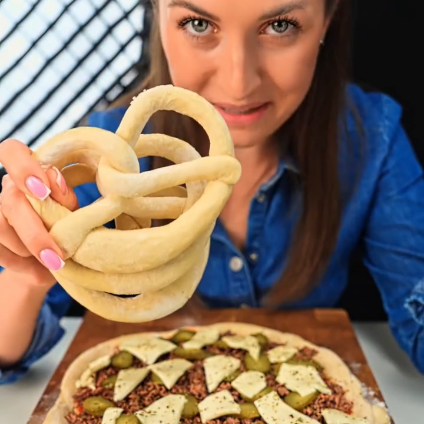 A woman in a blue shirt holds up a bundle of uncooked pretzels above a topped raw pizza on a wooden board, with a dark background and a window grid visible behind her.
