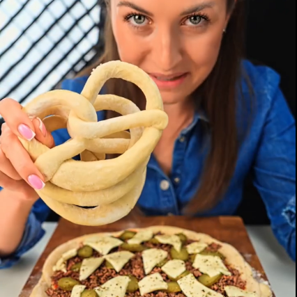 A woman in a blue shirt holds up a bundle of uncooked pretzels above a topped raw pizza on a wooden board, with a dark background and a window grid visible behind her.