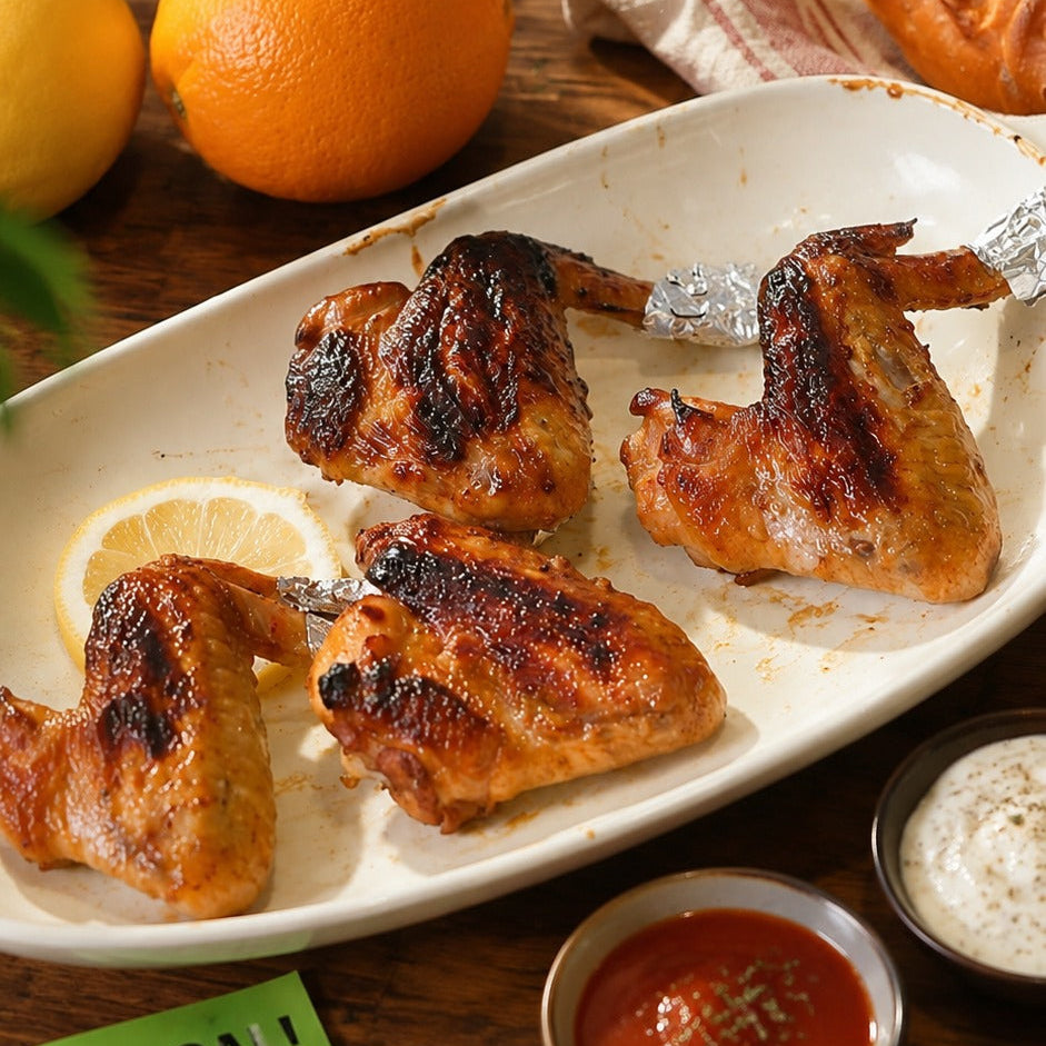Grilled chicken wings served on a white oval platter with lemon slices, set on a wooden table with soccer-themed props including a soccer ball, bread, and small dipping sauces.