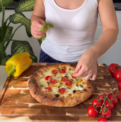 A woman in a white tank top is preparing a fresh pizza topped with cherry tomatoes, black olives, and green herbs on a wooden cutting board, with a yellow bell pepper and cherry tomatoes nearby in a kitchen setting.